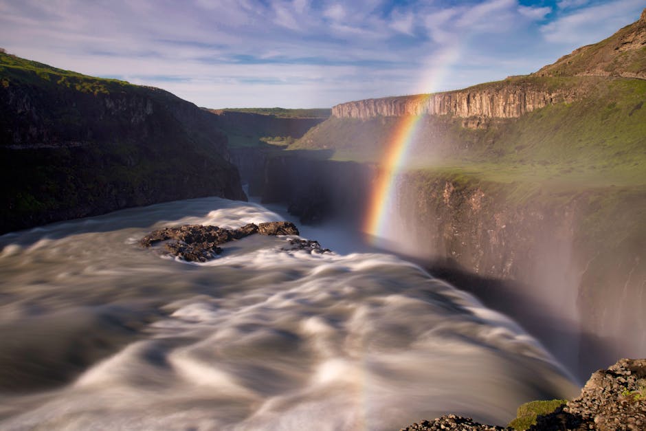 A wide view of a powerful river flowing through a deep canyon, with swirling, frothy white water in the foreground. Steep, rugged cliffs rise on both sides, covered in patches of green vegetation. A v
